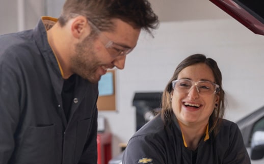 Two Smiling General Motors Certified Service Technicians in Safety Glasses Looking at a Laptop