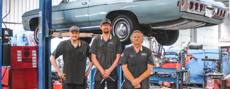 Three Auto Technicians Standing Under a Light Blue Classic Car on a Lift in a Service Bay