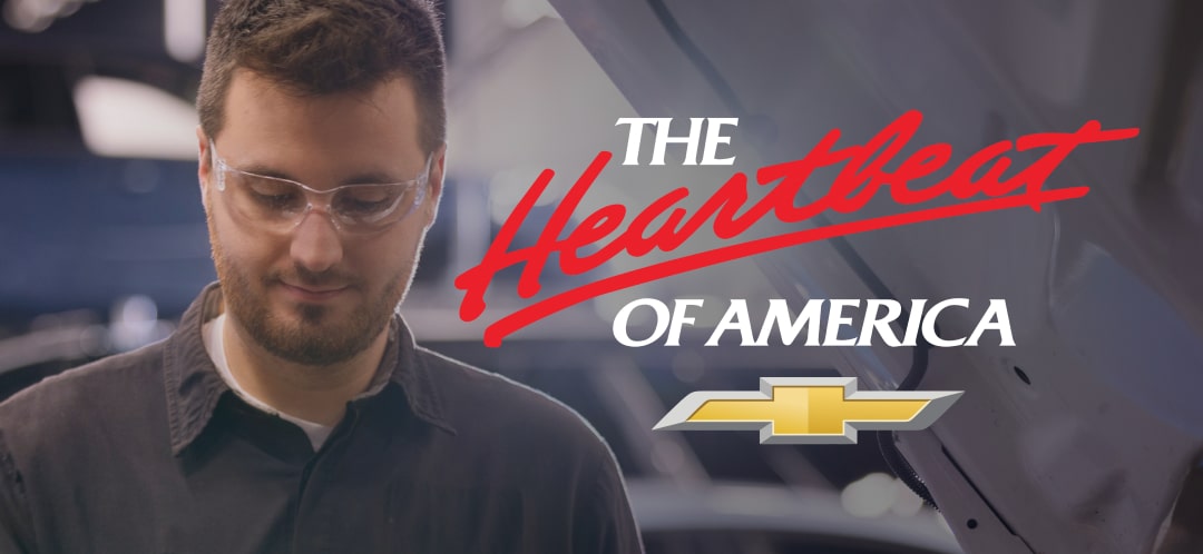 A Chevrolet Certified Service Technician Looks Downward with the Heartbeat of America Text and a Gold Chevrolet Logo Overlayed
