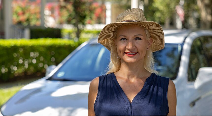 A woman in a hat and blue shirt smiling while standing in front of a white vehicle.