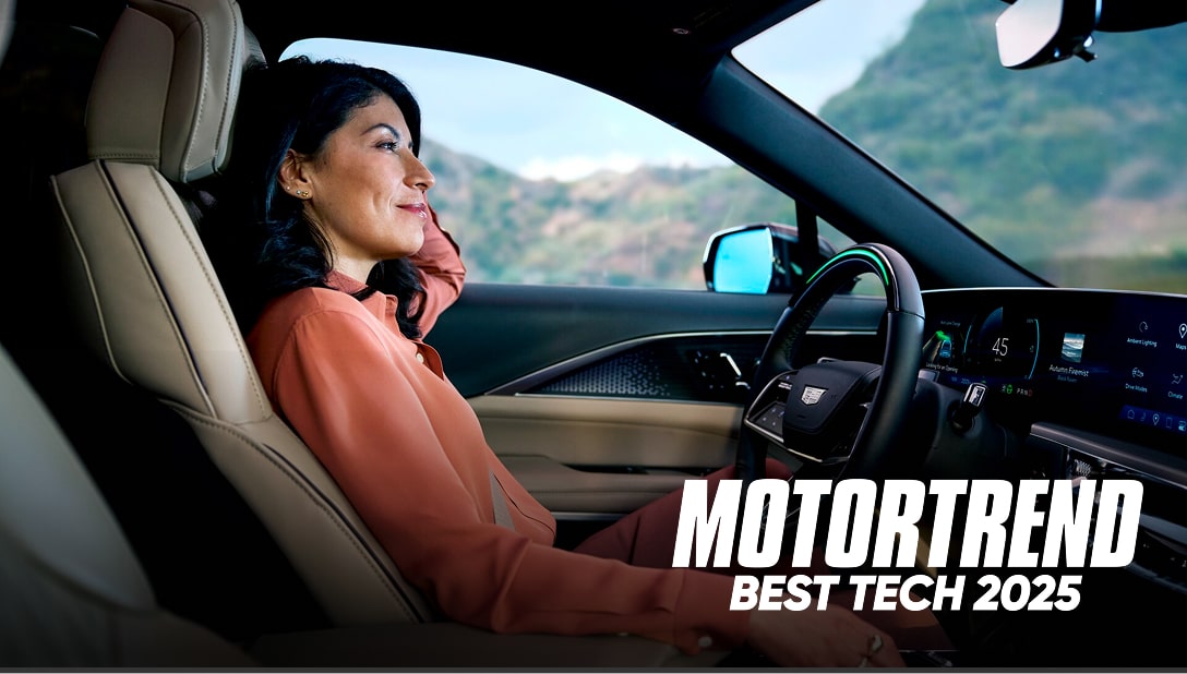 A woman with dark hair, wearing a reddish-orange shirt, is smiling while sitting in the driver seat of a car as she looks out the window, with the words "MOTORTREND BEST TECH 2025" overlaid on the image.