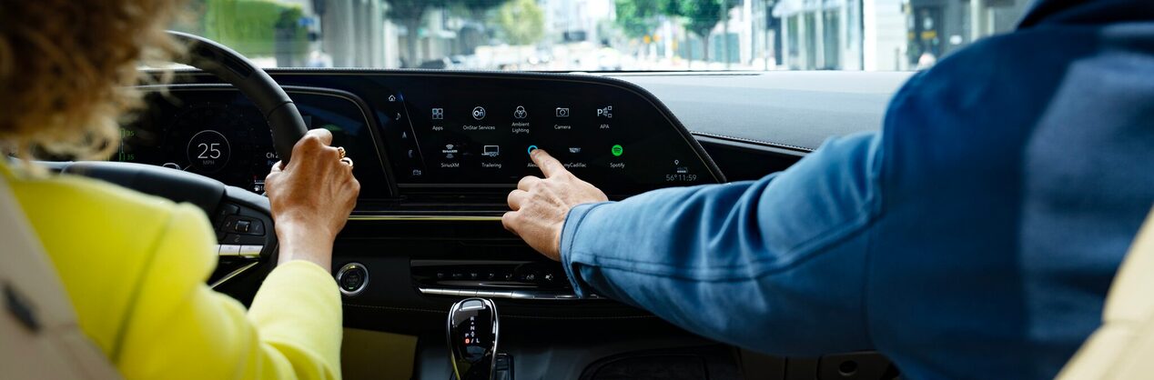 A woman in a yellow jacket holding the steering wheel and a man in a blue jacket pressing a button on the infotainment system