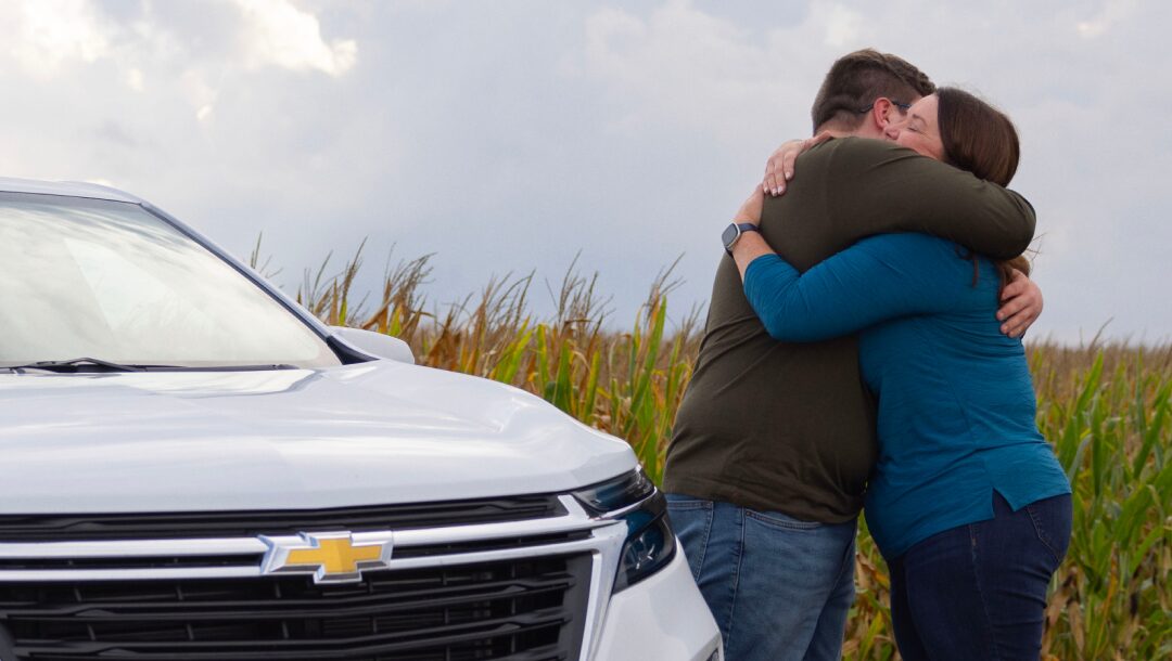 A man and woman embrace next to the grille of a white Chevrolet in a field of tall grass under an overcast sky.