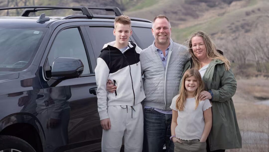 A smiling family of four stands together next to a dark-colored chevrolet SUV on a grassy hillside.