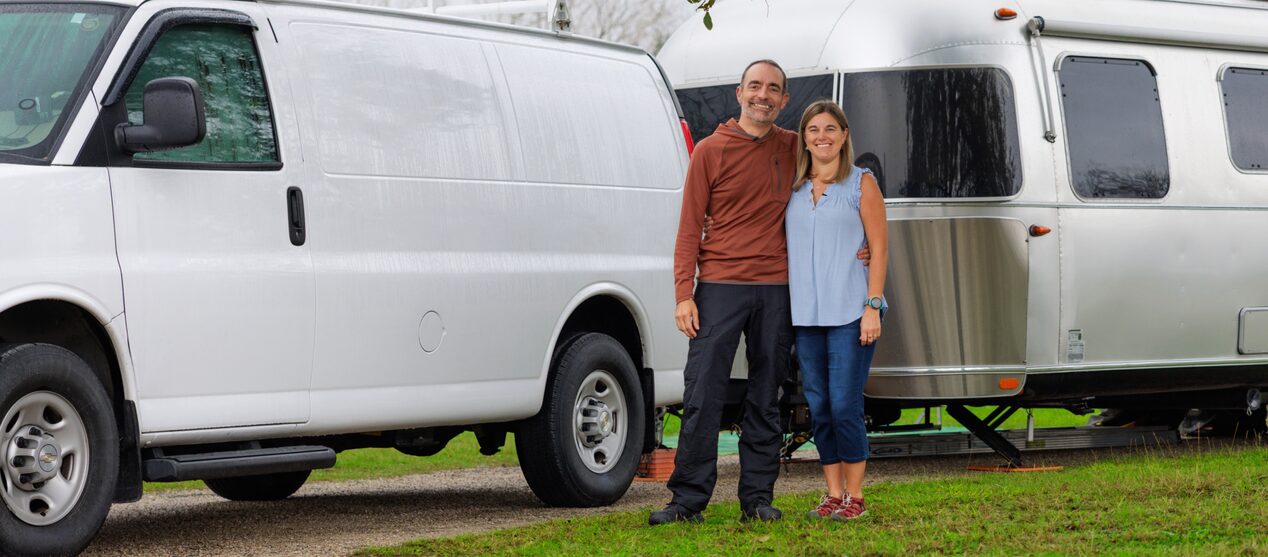 A man and woman stand in front of a white cargo van and a silver travel trailer on a grassy campground.
