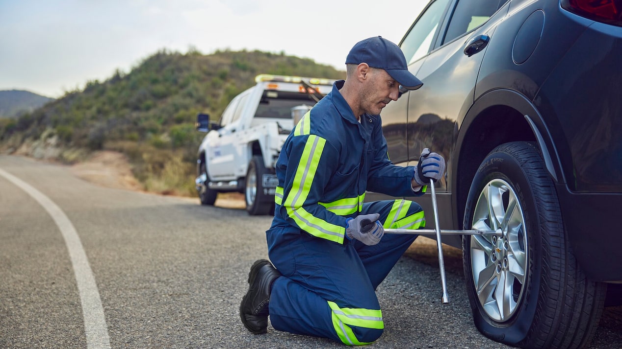 A roadside assistance technician in a blue jumpsuit with neon yellow stripes is kneeling on an asphalt road next to a dark vehicle, using a lug wrench to change a flat tire, with a white tow truck visible in the background.