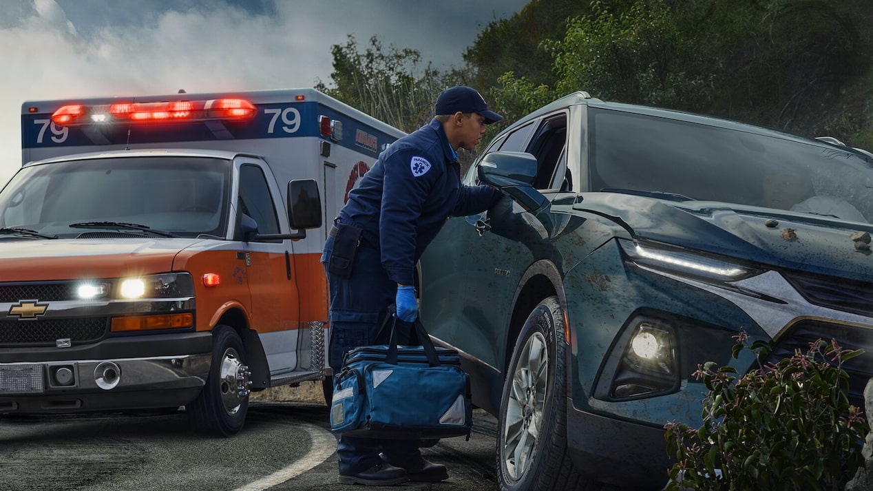 A first responder, wearing a uniform and carrying a medical bag, attends to a damaged dark SUV on the side of a road, with an ambulance with flashing lights visible just behind the vehicle.