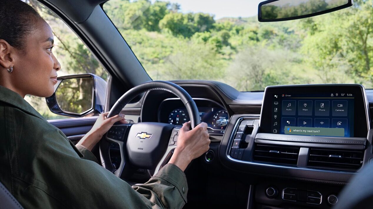 A person drives a Chevrolet vehicle, with the infotainment screen showing various apps like Google Assistant and Spotify.