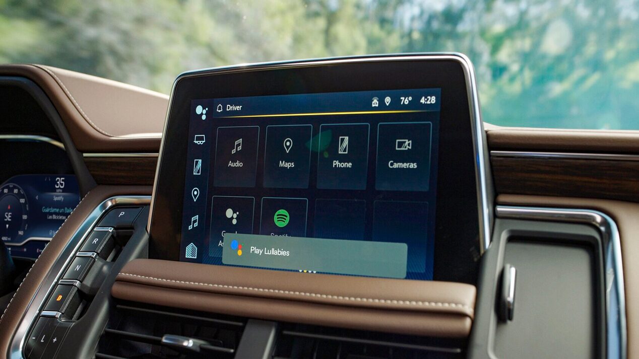 The vehicle's infotainment screen displays various apps and the Google Assistant search bar at the bottom, within a brown interior.