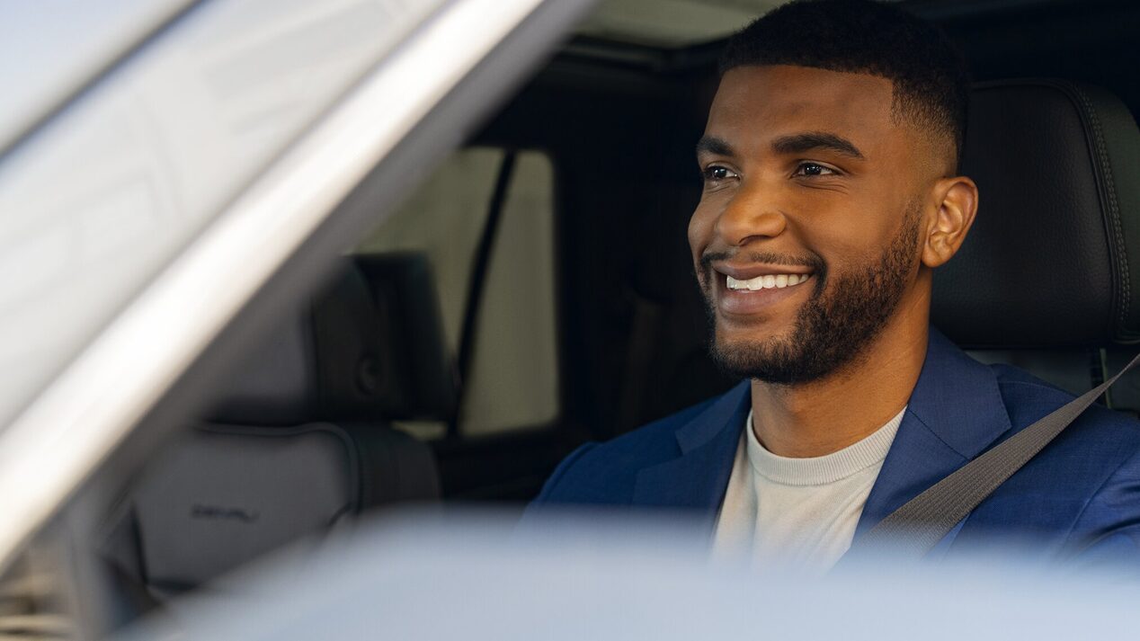 A smiling, well-dressed man wearing a seatbelt and a blue suit jacket is looking out of the driver's side window of a vehicle.