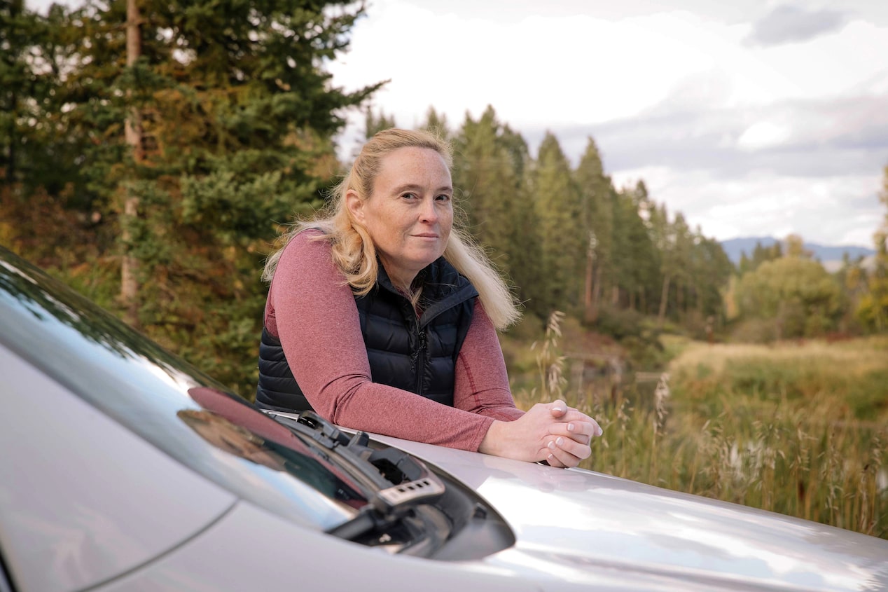 Woman Wearing Vest Leans on Front End of Vehicle