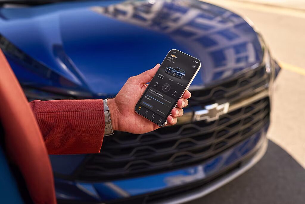 A person's left hand holding a smartphone displaying the OnStar app and a blue Chevy vehicle in the background.