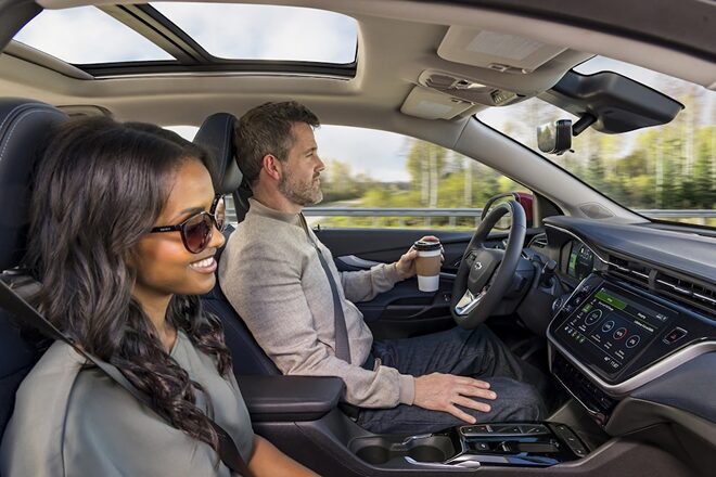 A man sitting in the driver's seat and holding a coffee with his left hand and a woman sitting in the passenger seat.